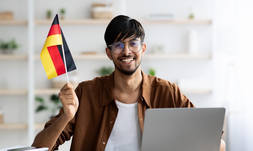 Smiling indian guy student showing flag of Germany, using laptop